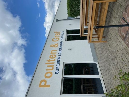 Das Bild zeigt das Poulten & Graf Firmengebäude. Im Vordergrund eine Holzbank auf der Terrasse, im Hintergrund blau-weißer Himmel.